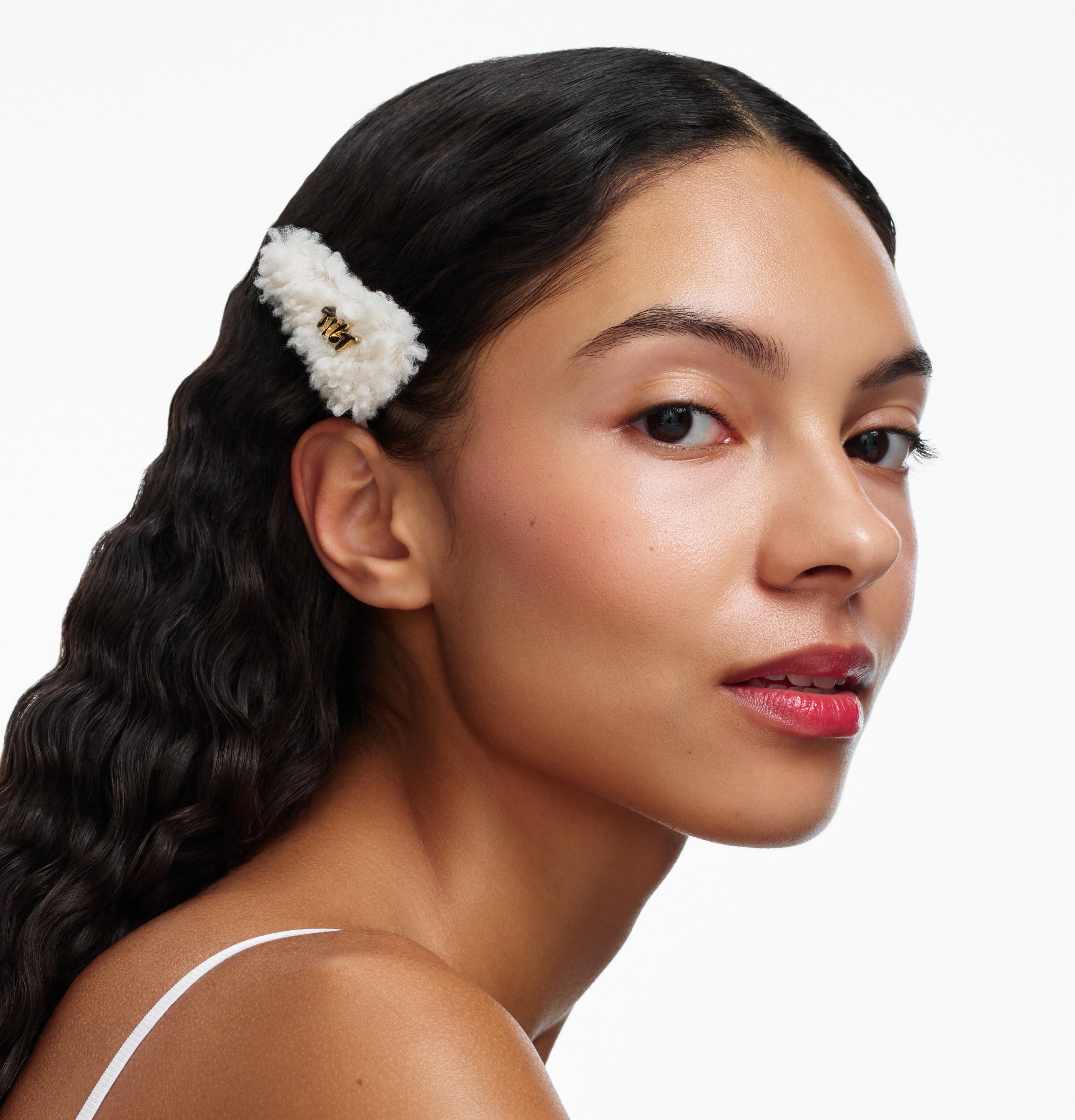 Person with long dark hair wearing a fluffy off-white hair clip and white top against a plain background.