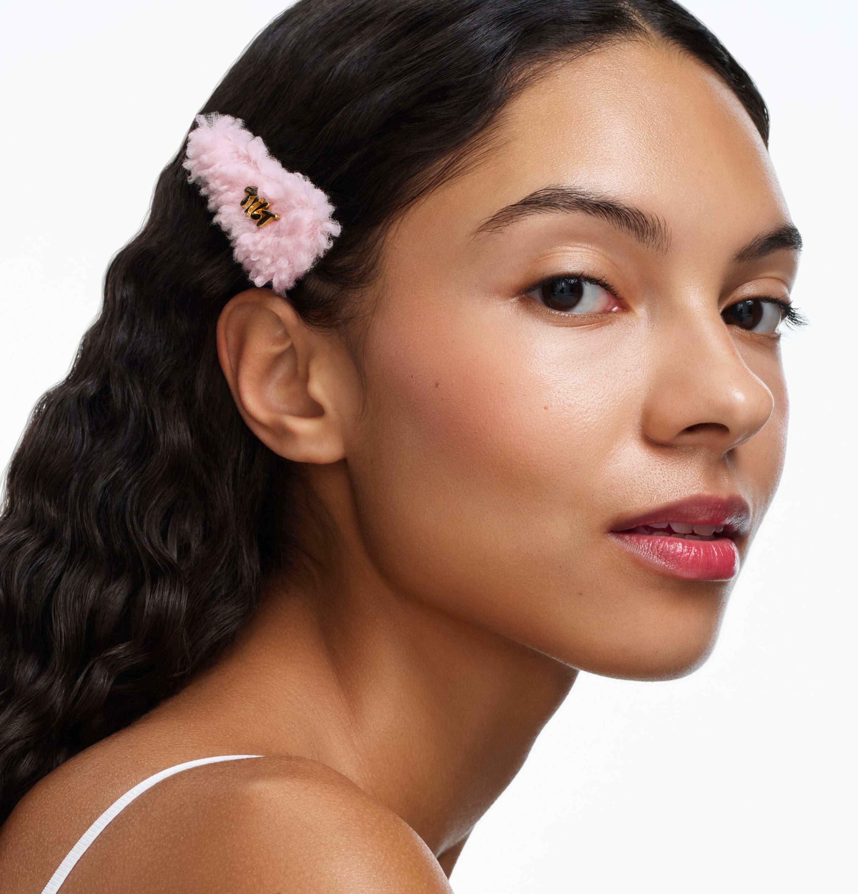 Person with long dark hair wearing a fluffy pink hair clip and white top against a plain background.