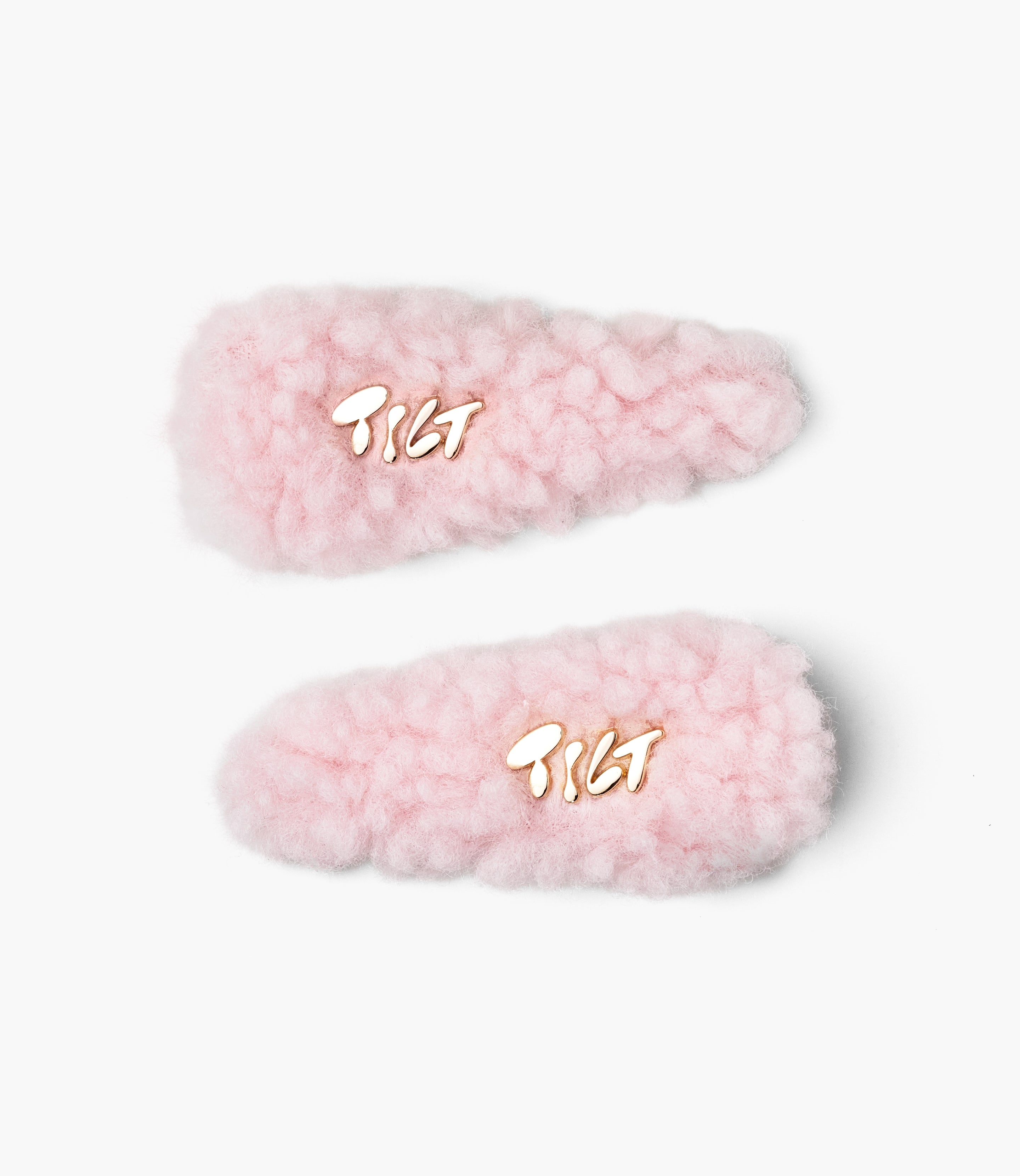 Two pink fluffy hair clips that say 'Tilt' in gold lettering against a plain background.