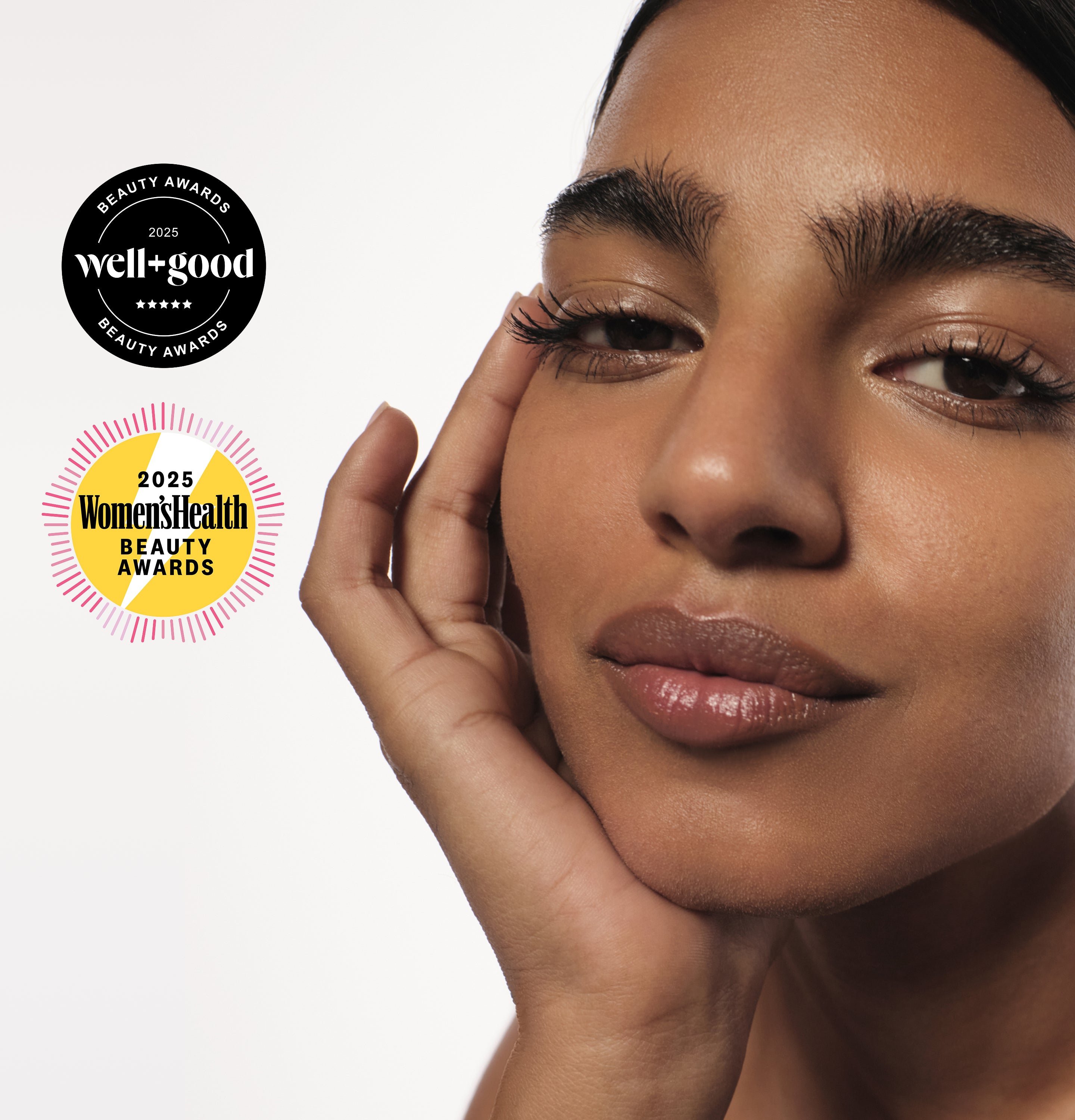 Close-up of a woman with glowing skin and defined brows, resting her face on her hand. 2025 Well+Good and Women’s Health Beauty Awards badges appear beside her.