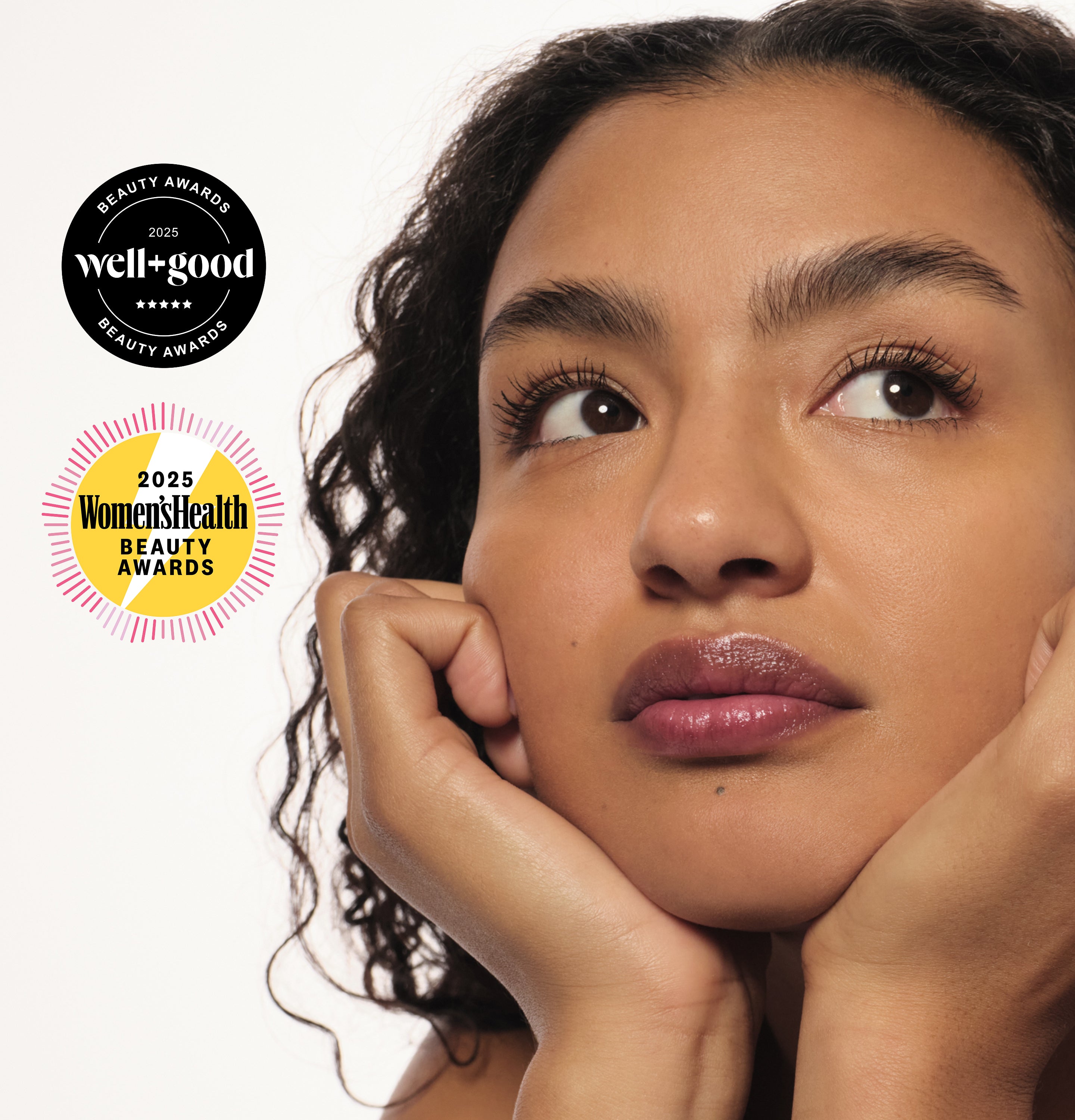 Woman with glossy lips rests her chin on her hands, looking upward. 2025 Well+Good and Women’s Health Beauty Awards badges appear beside her.